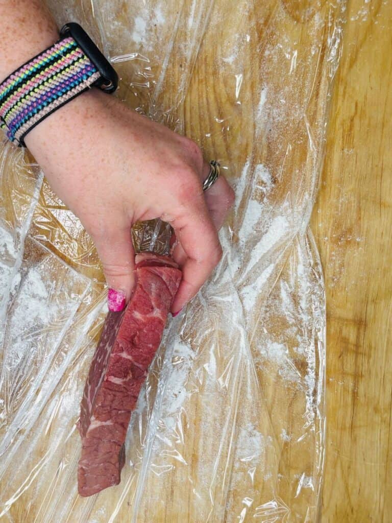 A hand with a colorful wristband holds a raw steak on a flour-dusted wooden surface covered with plastic wrap.