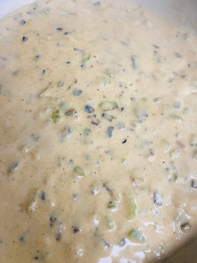 Creamy beige soup or sauce with visible chunks of vegetables and herbs, including green and brown pieces, simmering in a pot.