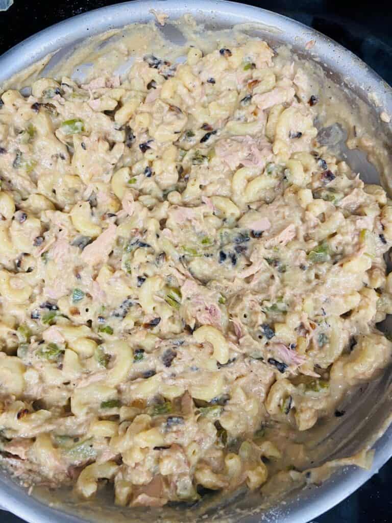 A close-up of a creamy macaroni tuna casserole in a metal pan, with elbow pasta, chunks of tuna, black olives, and green vegetables mixed together.