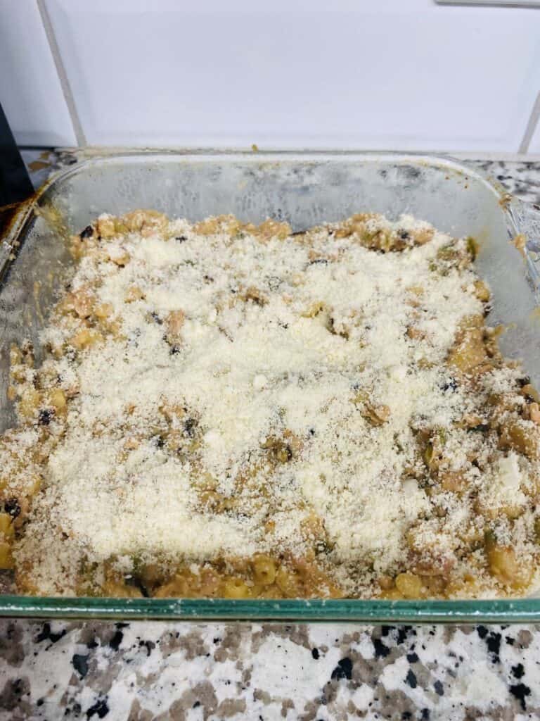 A glass baking dish filled with a baked casserole, topped with a layer of grated cheese or breadcrumbs, sits on a granite countertop in a kitchen with white tile backsplash.