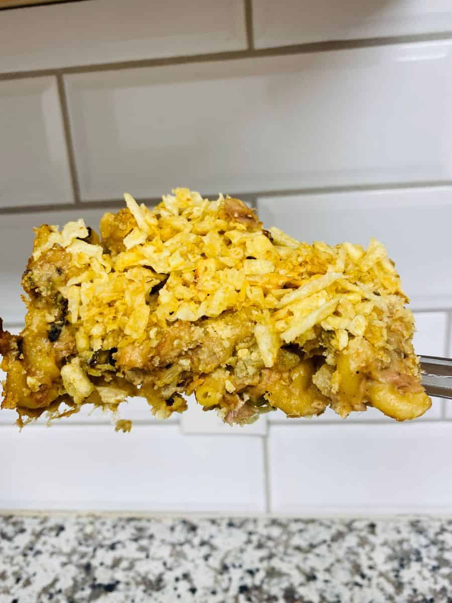A fork holds a serving of casserole topped with crushed chips, with a tiled backsplash and speckled countertop in the background.
