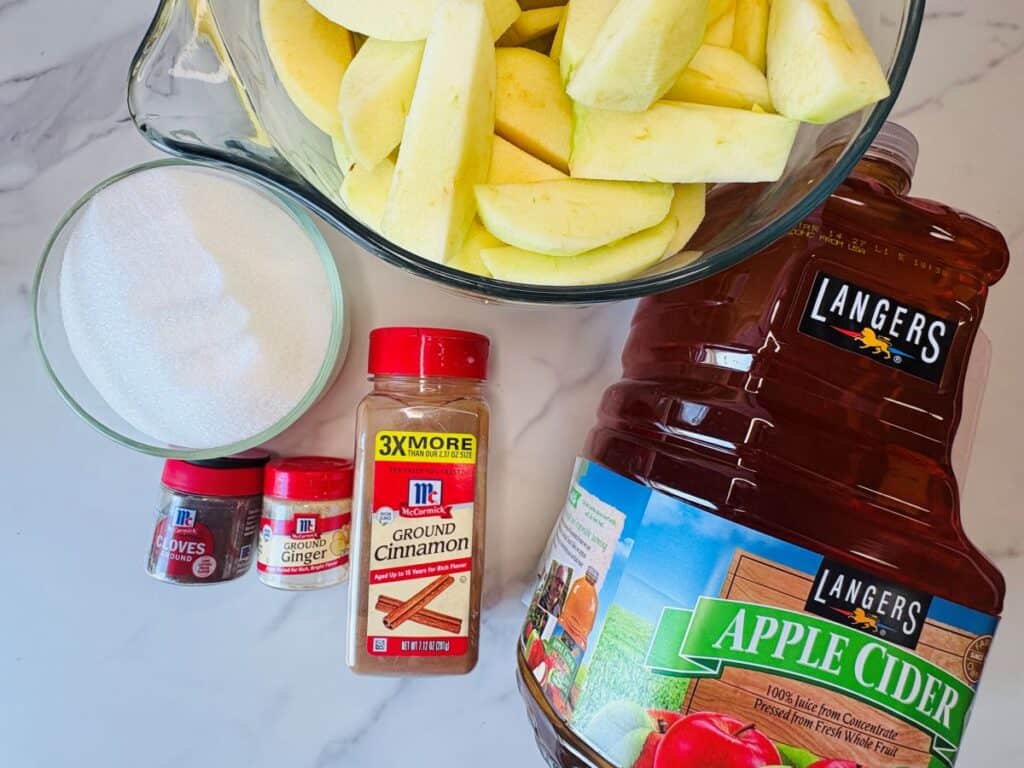 A bowl of sliced apples, a jar of apple cider, a bowl of sugar, and small containers of ground cloves, ground ginger, and ground cinnamon are arranged on a white marble surface.