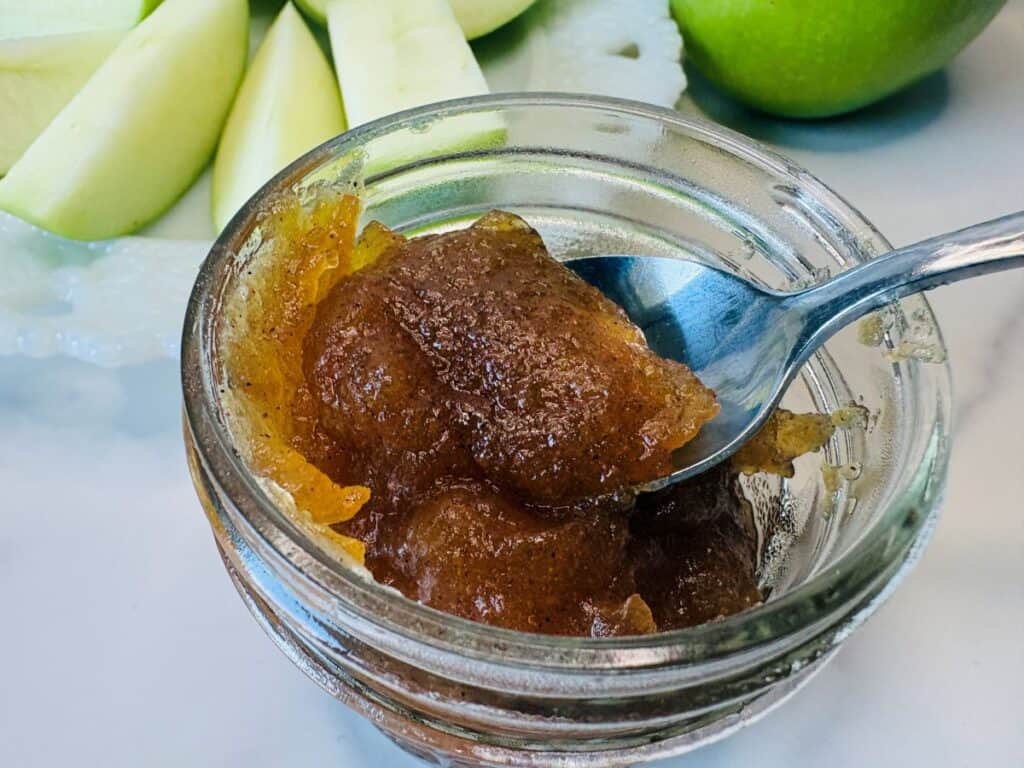 A spoon scoops a thick, brown, cinnamon-spiced apple butter from a small glass jar. Sliced green apples and a whole green apple are visible in the background.