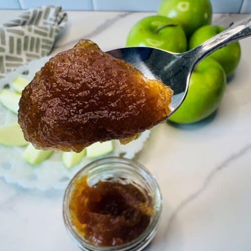 A spoonful of brown apple butter is held above a jar, with green apples and slices on a plate in the background on a marble countertop.