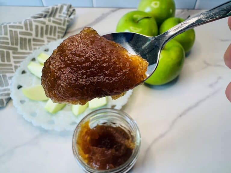 A spoonful of brown apple butter is held above a jar, with green apples and slices on a plate in the background on a marble countertop.