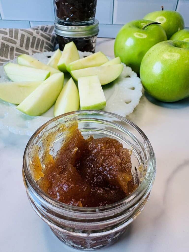 Open jar of apple butter in the foreground, a plate with sliced green apples, whole green apples, and more jars in the background on a white countertop.