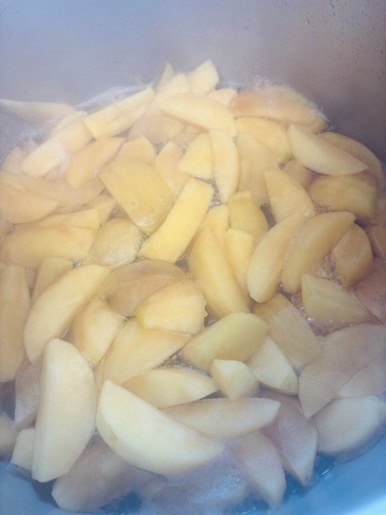 Sliced yellow apples simmering in a pot, with steam rising and some liquid visible among the fruit pieces.