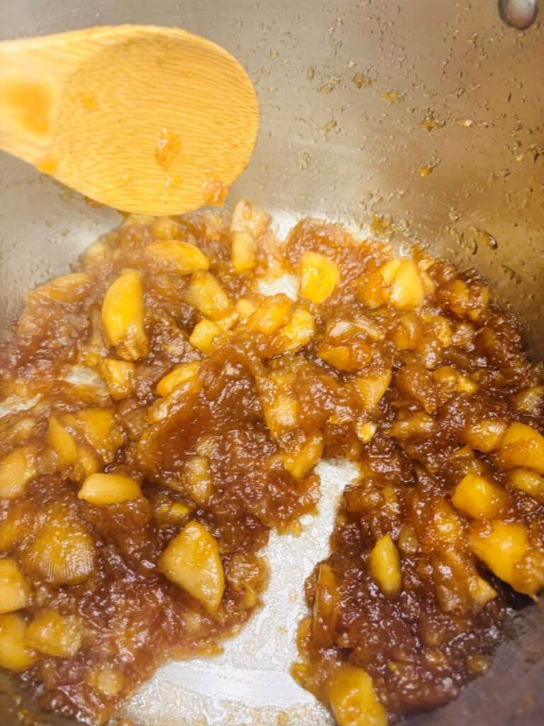 Chopped apples cooking in a pot with sugar and spices, forming a caramelized, chunky mixture. A wooden spoon is stirring the contents.