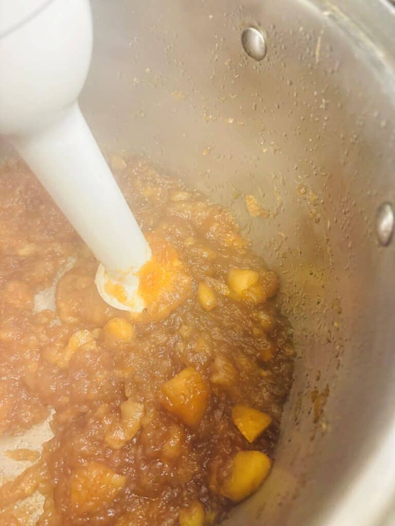 A hand blender blends chunky apple and brown mixture in a large metal pot, possibly making applesauce or apple butter. Steam rises, and the sides of the pot show condensation and food splatters.