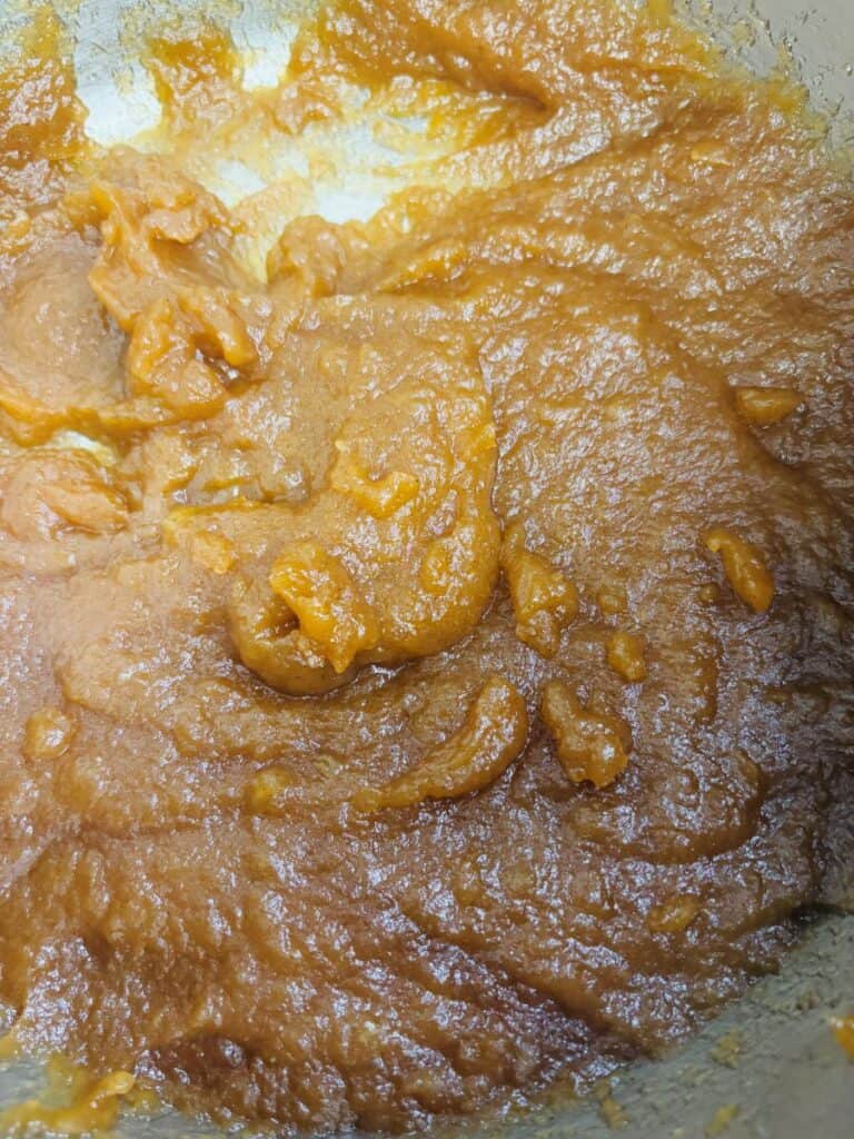 Thick, orange-brown paste with a glossy texture, spread unevenly in a metal bowl, possibly a cooked fruit puree or sweet mixture.