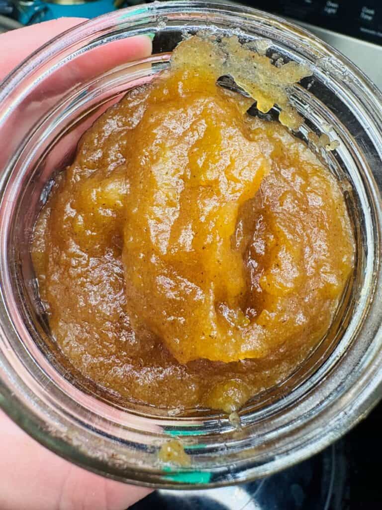 A close-up of a glass jar filled with a thick, brownish, grainy paste, possibly homemade apple butter or fruit preserve, with some residue near the rim.