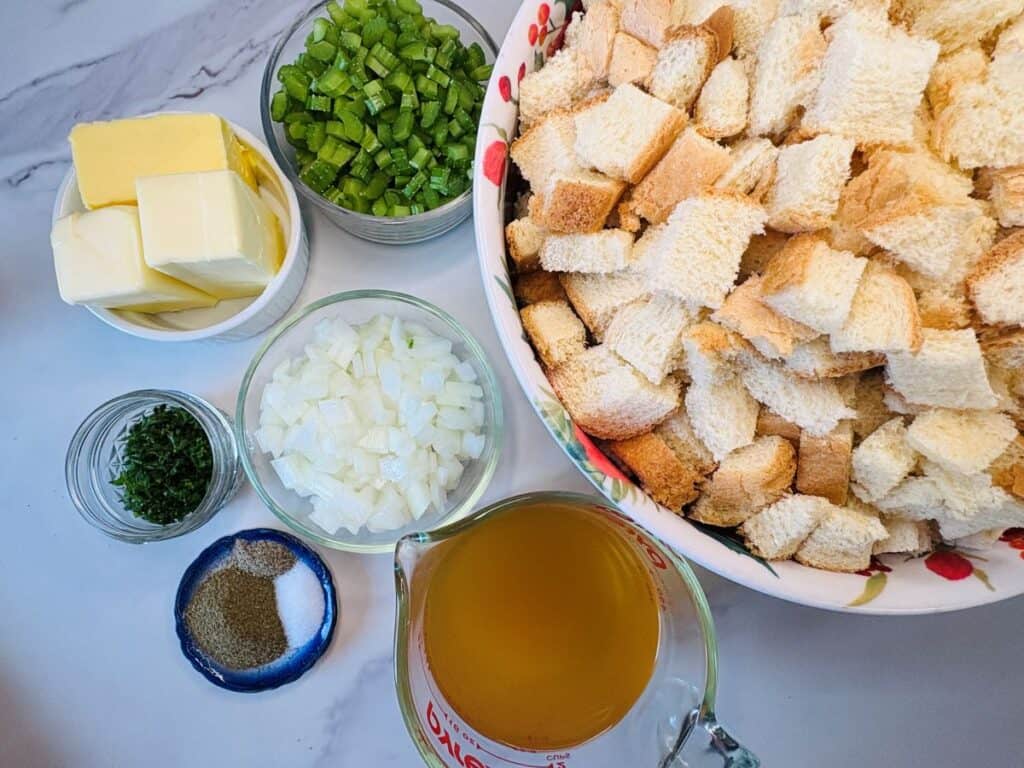 Bowls with cubed bread, diced onions, chopped celery, parsley, butter sticks, salt, pepper, and a measuring cup of broth on a marble surface, arranged for preparing a stuffing recipe.