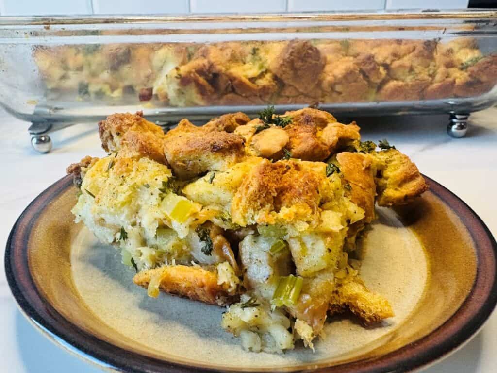 A plate with a serving of golden brown bread stuffing featuring celery and herbs, with a glass baking dish containing more stuffing in the background.