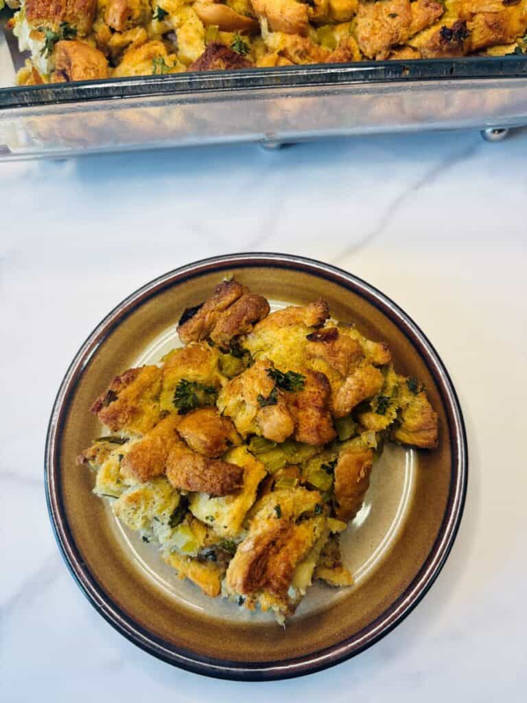 A plate with a serving of bread stuffing, showing golden, crispy bread cubes mixed with herbs and vegetables. The rest of the stuffing is visible in a baking dish in the background.