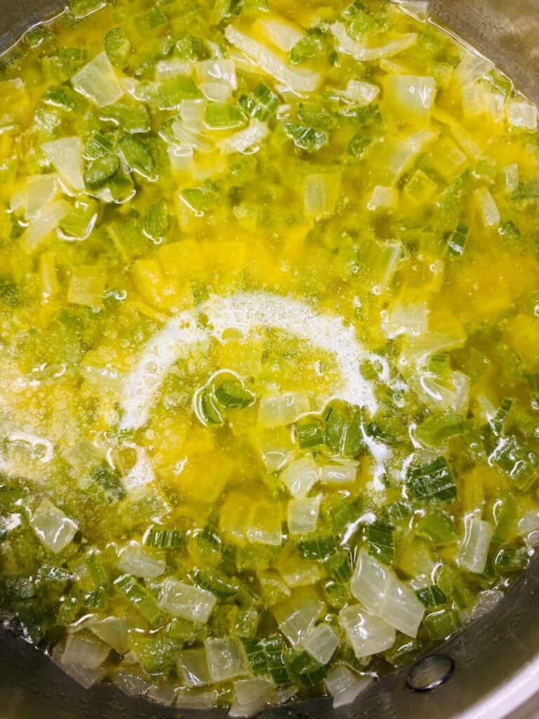Diced onions and celery sautéing in melted butter in a pot, with visible bubbles and a glossy surface.