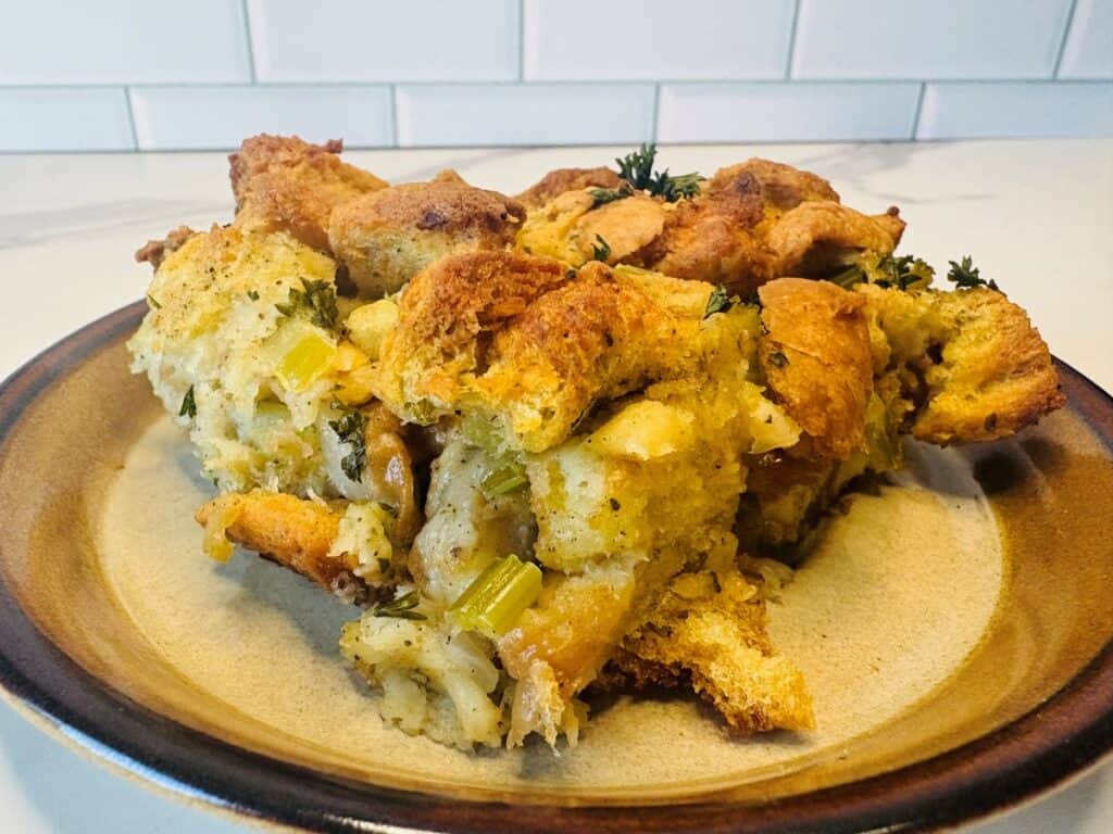 A slice of golden-brown bread stuffing with herbs and celery sits on a tan plate, against a white tile background.