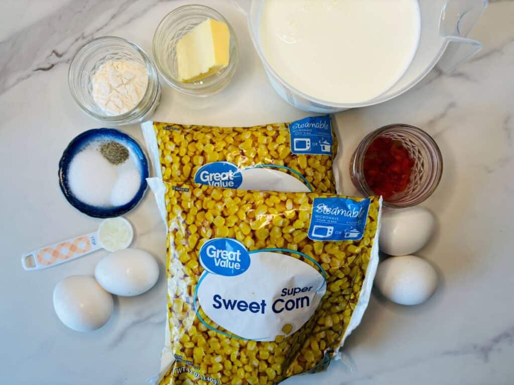 Ingredients for a recipe displayed on a marble surface: two bags of frozen sweet corn, three eggs, milk, flour, butter, salt, pepper, sugar, and a small bowl of red diced peppers.