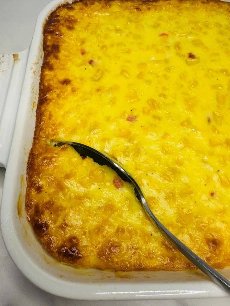 A golden-brown baked corn casserole in a white dish, with a spoon scooping out a portion. The surface looks cheesy and slightly crispy around the edges.