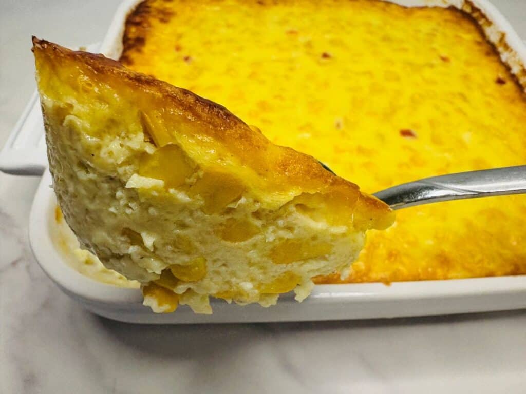 A close-up of a serving of creamy baked corn casserole on a spoon, held above a white baking dish filled with more casserole.