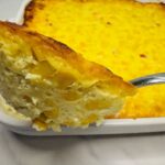 A close-up of a serving of creamy baked corn casserole on a spoon, held above a white baking dish filled with more casserole.