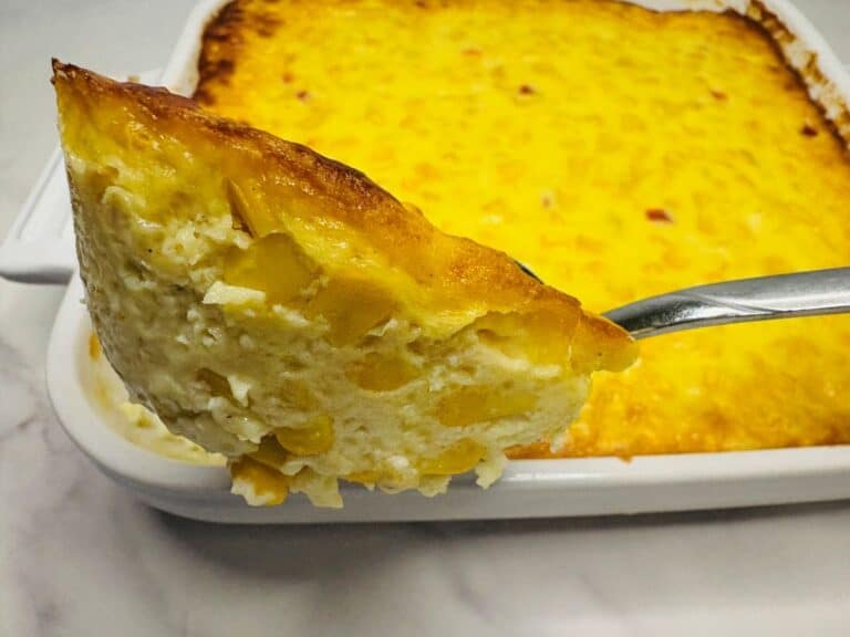 A close-up of a serving of creamy baked corn casserole on a spoon, held above a white baking dish filled with more casserole.