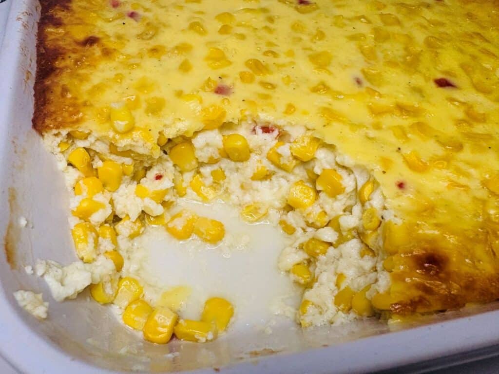 A baked corn casserole in a white dish, with a portion scooped out showing creamy corn filling and a golden, slightly browned top.