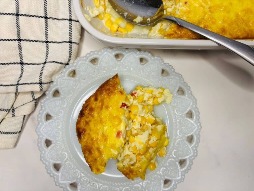 A slice of cheesy corn casserole on a white decorative plate, with a serving dish and spoon nearby, and a checkered towel to the side.
