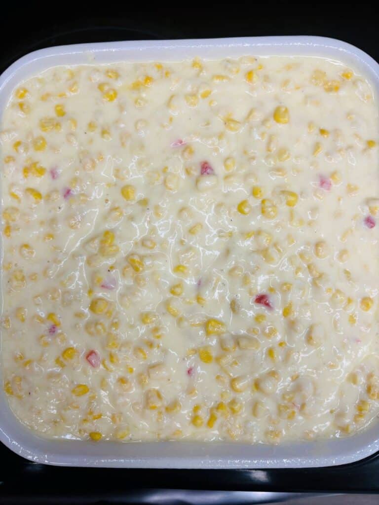 A close-up of a creamy corn casserole mixture with visible yellow corn kernels and small bits of red pepper in a white square dish.