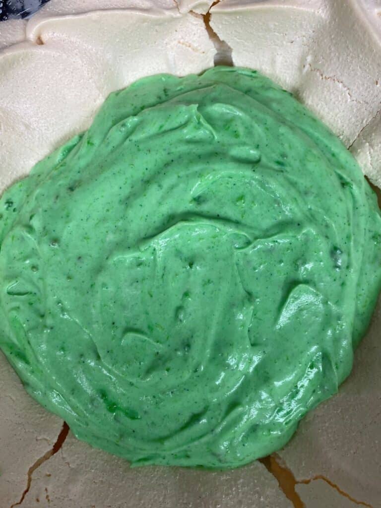 A close-up of a cracked, beige meringue base filled with a thick layer of glossy, mint green cream or mousse in the center.