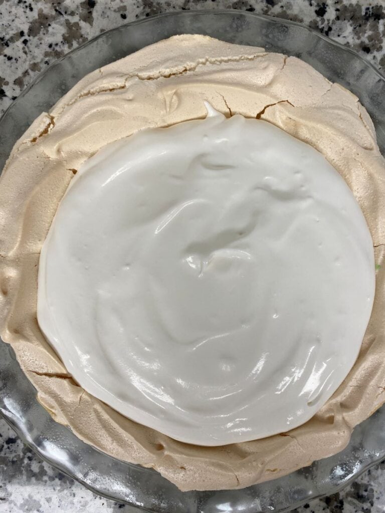 A round pavlova with a crisp, light tan meringue shell and a thick layer of glossy white whipped cream on top, sitting on a clear glass plate on a speckled countertop.