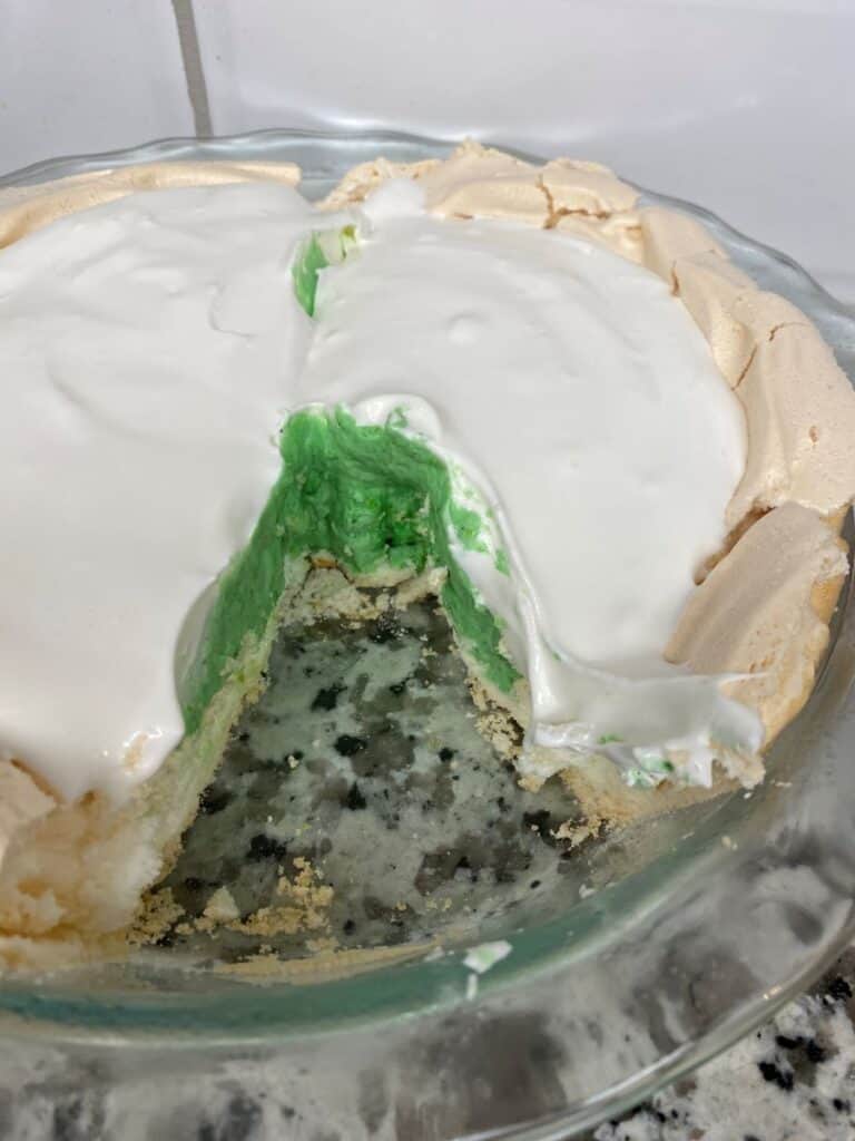 A pie with a white meringue topping and green filling, with a slice missing, sits in a glass dish on a granite countertop.