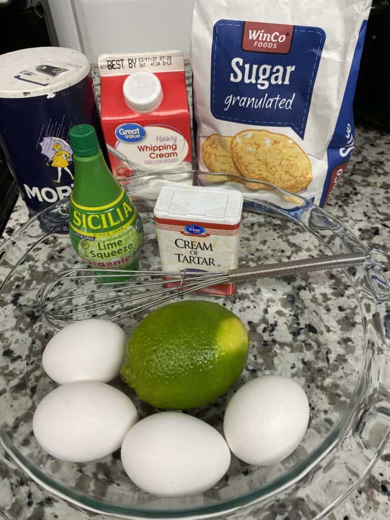 A bowl with four eggs, a lime, and a whisk sits on a counter surrounded by salt, lime juice, cream of tartar, heavy whipping cream, and sugar.