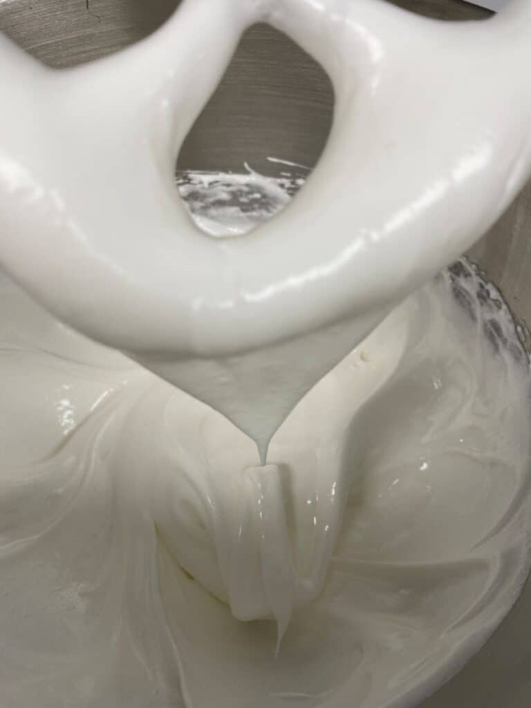 Thick, glossy white icing or meringue is being mixed in a bowl with a stand mixer attachment, with peaks forming as it is lifted.