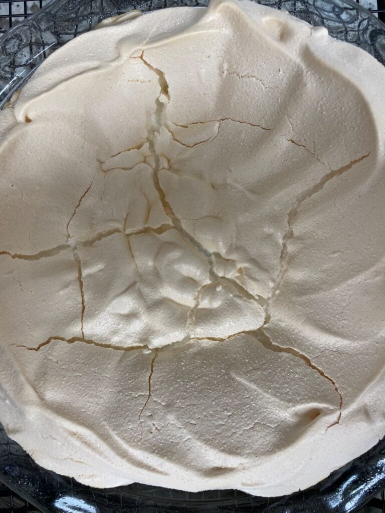 A round, baked meringue with a crisp, cracked surface sits on a glass plate. The meringue is pale and slightly golden around the edges.