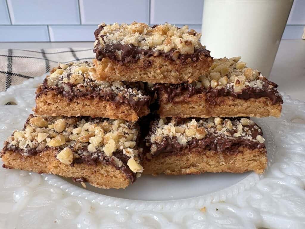 Five nut-topped chocolate bars stacked on a white decorative plate, with a glass of milk and checkered napkin in the background.