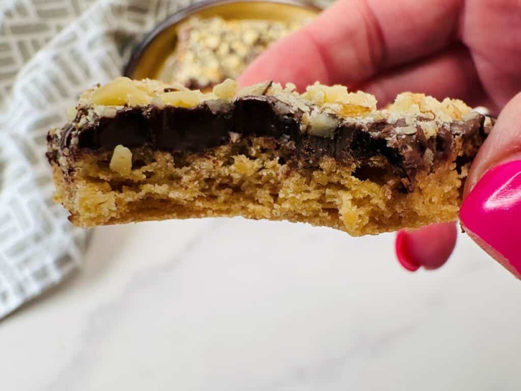 A hand with pink nail polish holds a half-eaten baked cookie bar topped with chocolate chips and nuts, with a blurred cookie in the background.