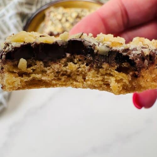 A hand with pink nail polish holds a half-eaten baked cookie bar topped with chocolate chips and nuts, with a blurred cookie in the background.