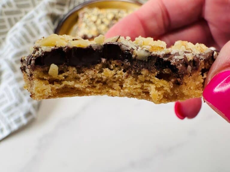 A hand with pink nail polish holds a half-eaten baked cookie bar topped with chocolate chips and nuts, with a blurred cookie in the background.