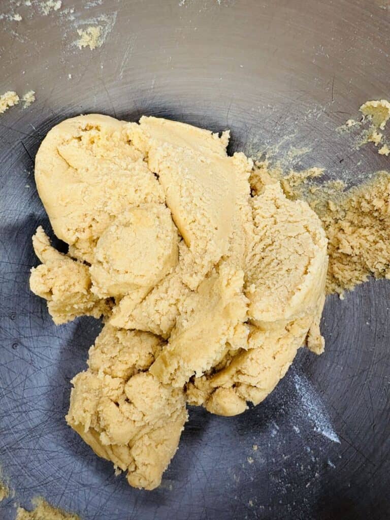 A close-up of pale yellow cookie dough in a metal mixing bowl, with some dough stuck to the sides of the bowl.