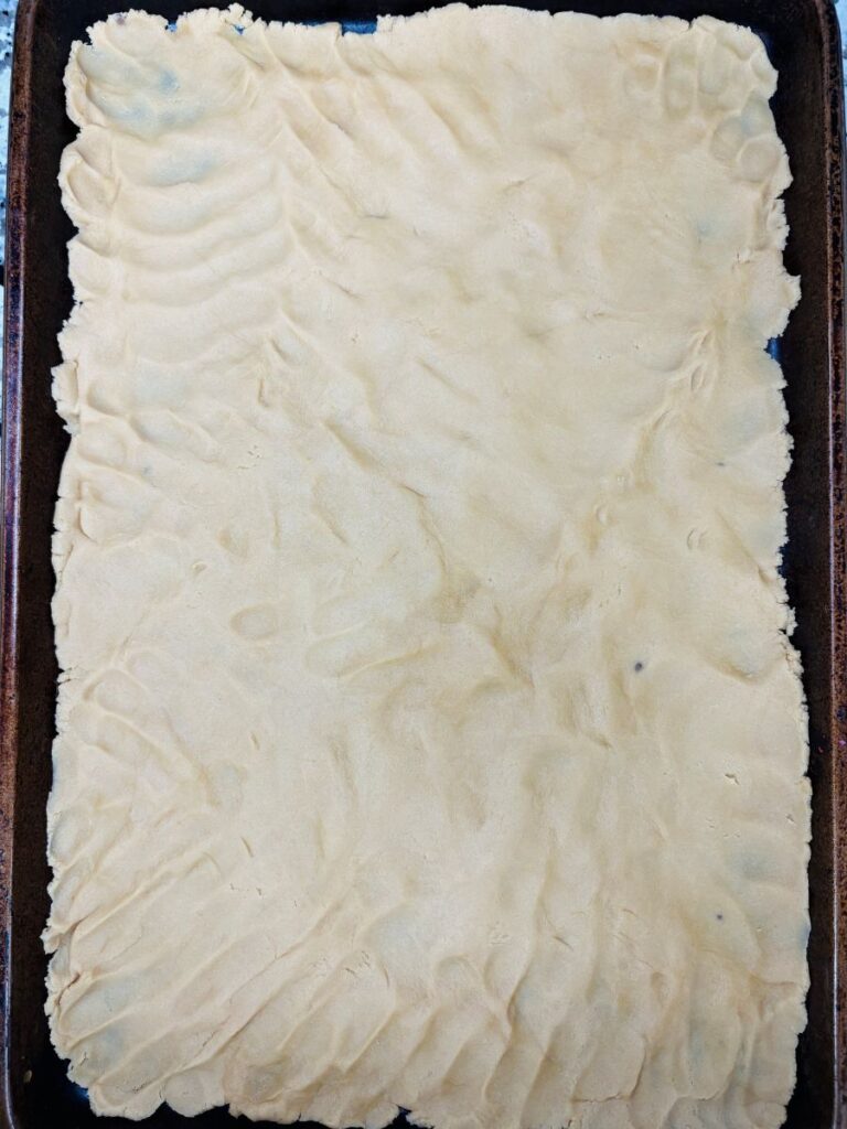 A baking sheet covered with an even layer of unbaked dough, showing finger marks from spreading.