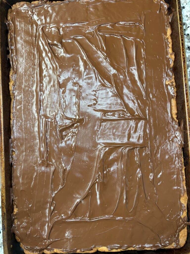 A rectangular baking tray filled with a dessert topped with a smooth layer of melted chocolate, covering the entire surface. The chocolate has visible spreading marks.