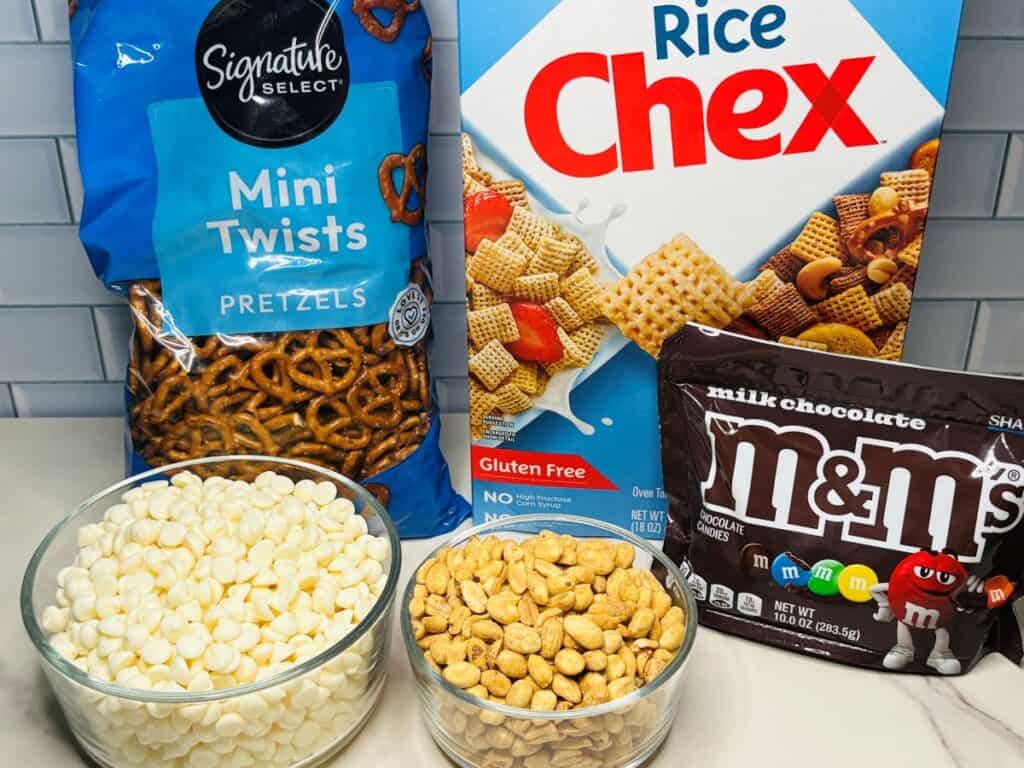 A bag of pretzels, a box of Rice Chex, a bag of MMs, a bowl of white chocolate chips, and a bowl of peanuts are arranged on a kitchen counter.
