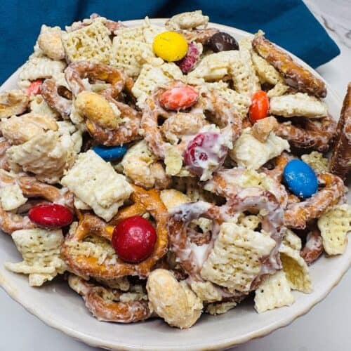 A bowl filled with a snack mix of pretzels, Chex cereal, peanuts, colorful chocolate candies, and a white coating, set on a marble surface with a blue cloth in the background.