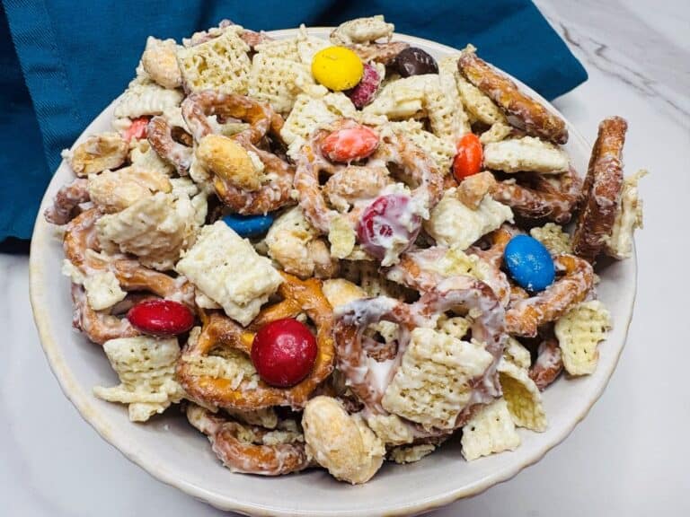 A bowl filled with a snack mix of pretzels, Chex cereal, peanuts, colorful chocolate candies, and a white coating, set on a marble surface with a blue cloth in the background.