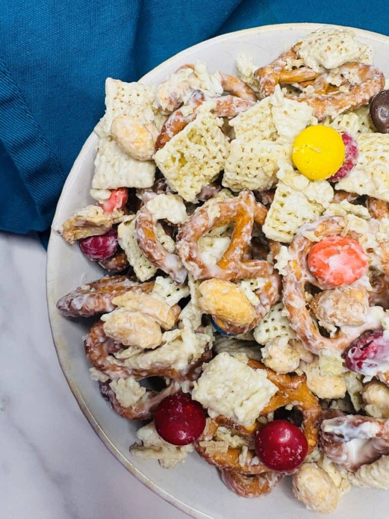 A close-up of a snack mix with pretzels, Chex cereal, red and yellow chocolate candies, and nuts, all coated in a white glaze, in a white bowl next to a blue cloth.