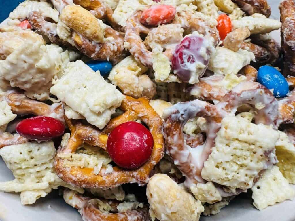 A close-up of a snack mix with pretzels, cereal squares, candy-coated chocolates, and nuts covered in a white coating.