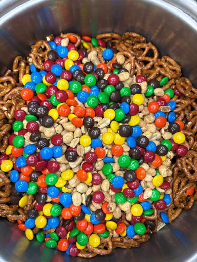 A stainless steel bowl filled with a snack mix of pretzels, peanuts, and colorful candy-coated chocolate pieces, such as MMs.