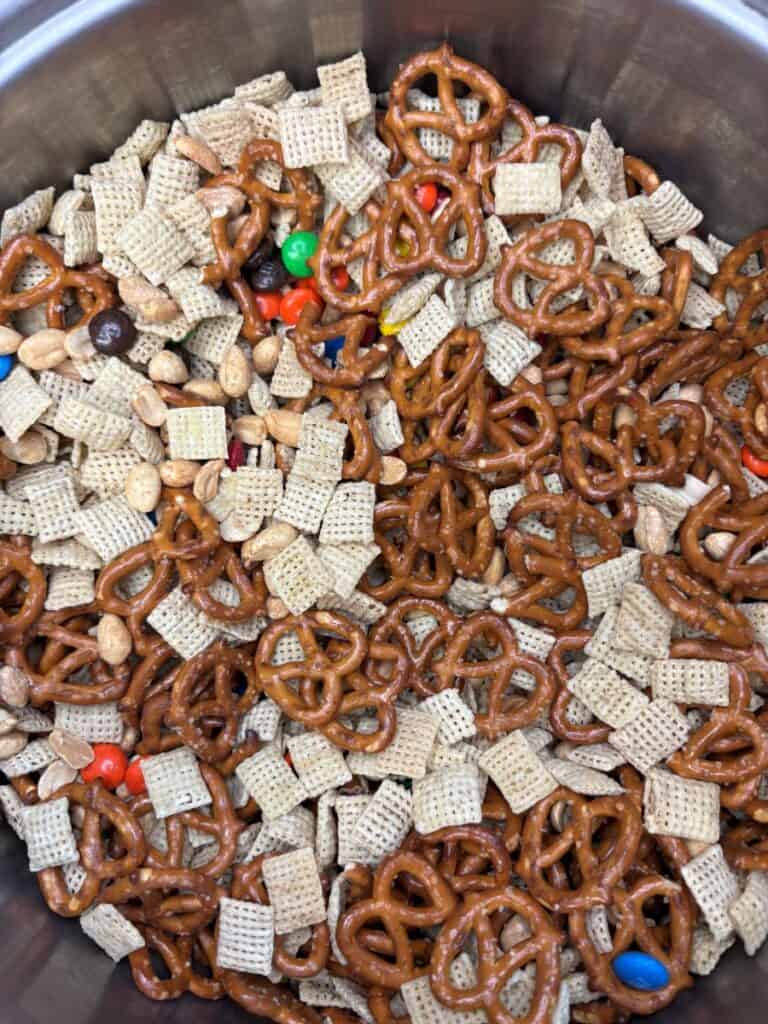 A bowl filled with small pretzels, Chex cereal squares, peanuts, and colorful candy-coated chocolates, creating a homemade snack mix.