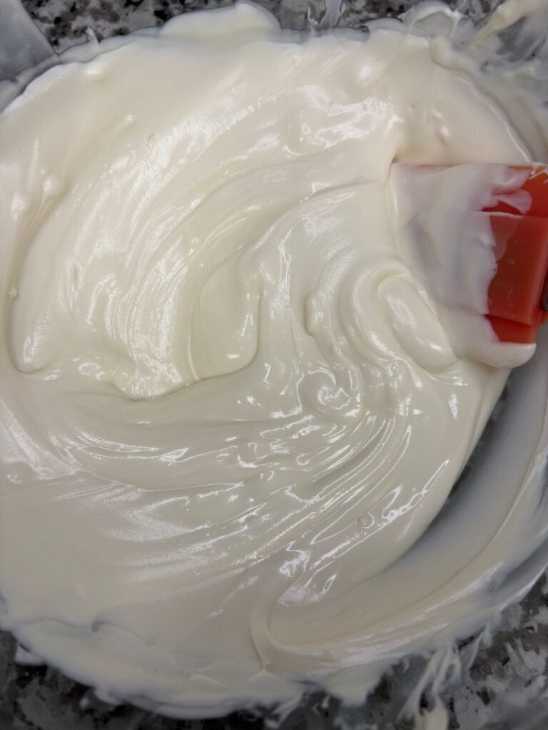 A close-up of a bowl filled with smooth, glossy white cream being mixed with an orange spatula.