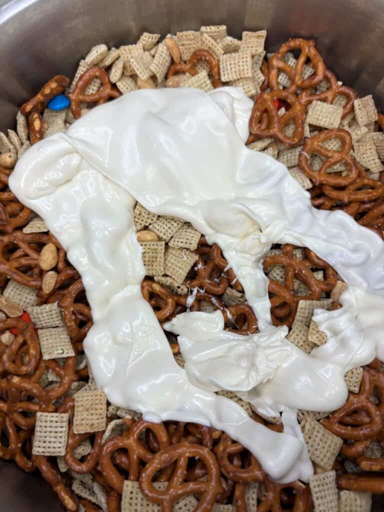 A close-up of a bowl containing pretzels, Chex cereal, peanuts, colorful candy pieces, and a layer of melted white chocolate on top.